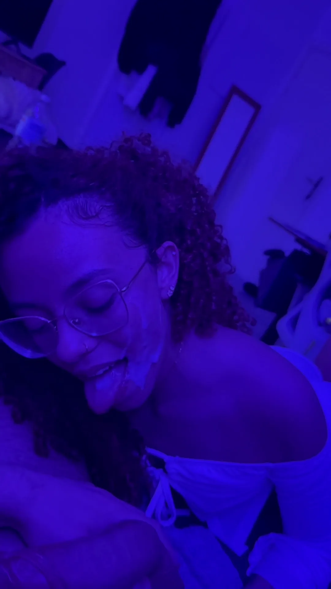 Goldencurls3 frame at 00:04, A smirk dances behind those mischief-perched glasses as she lounges in a cluttered, plant-adjacent sanctuary